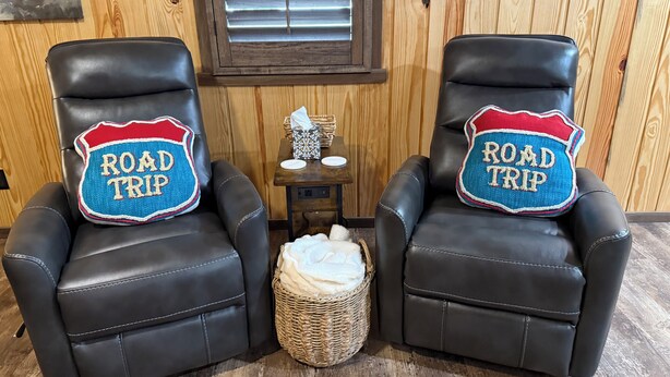 Two black leather chairs with 'ROAD TRIP' pillows, a small table with a tissue box, and a wicker basket on a wooden floor. - 66 Tiny House (Clinton)