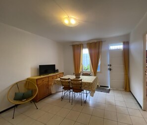 A small, clean room with a dining table, chairs, a television, and a chair with a cushion.