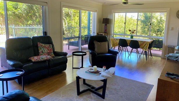 Ein geräumiges Wohnzimmer mit einer schwarzen Ledersofa, einem runden Couchtisch und einem Essbereich mit Tisch und Stühlen. Große Fenster bieten einen Ausblick ins Freie.