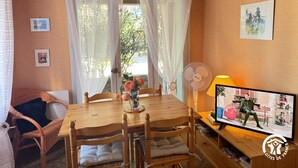 A dining area with a wooden table, chairs, a vase with flowers, a fan, and a television displaying an advertisement. - Pause Colliourencque (COLLIOURE)