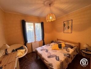 A bedroom with a bed, a chair, a table, a window, and a hanging light fixture.
