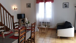 A dining area with a wooden table set for four, a TV on a shelf, and a window with sheer curtains.
