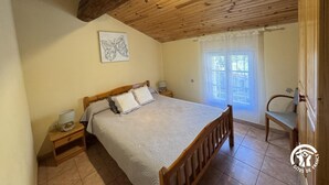A bedroom with a wooden ceiling, a bed with a patterned bedspread, a bedside table with a lamp, and a window with curtains.