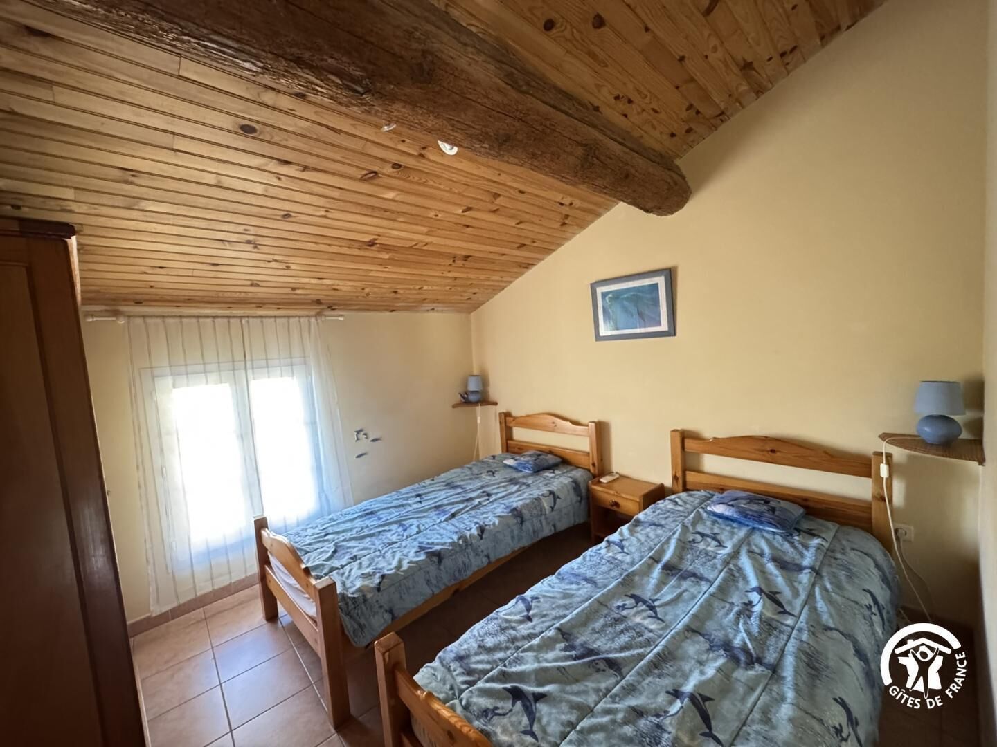 A small, cozy room with two wooden beds, a window with blinds, and a wooden ceiling.