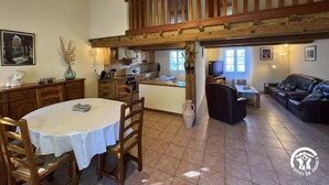 A spacious living area with a dining table, chairs, a sofa, and a TV. There is a kitchen area with cabinets and a window.