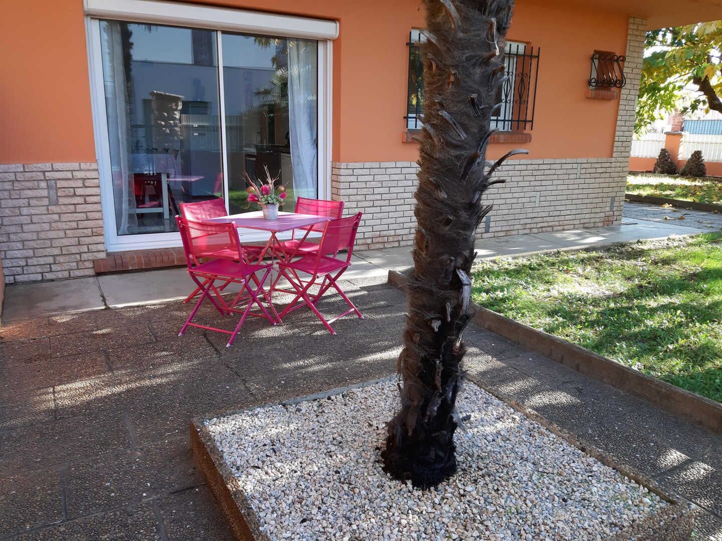 Eine Terrasse im Freien mit Tisch und Stühlen, einer Palme und einem Gebäude mit Backsteinfundament und orangenen Wänden.