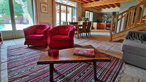 Een ruime woonkamer met een hoge plafond en zichtbare houten balken, met twee rode fauteuils, een salontafel met een boek en een grijze bank.