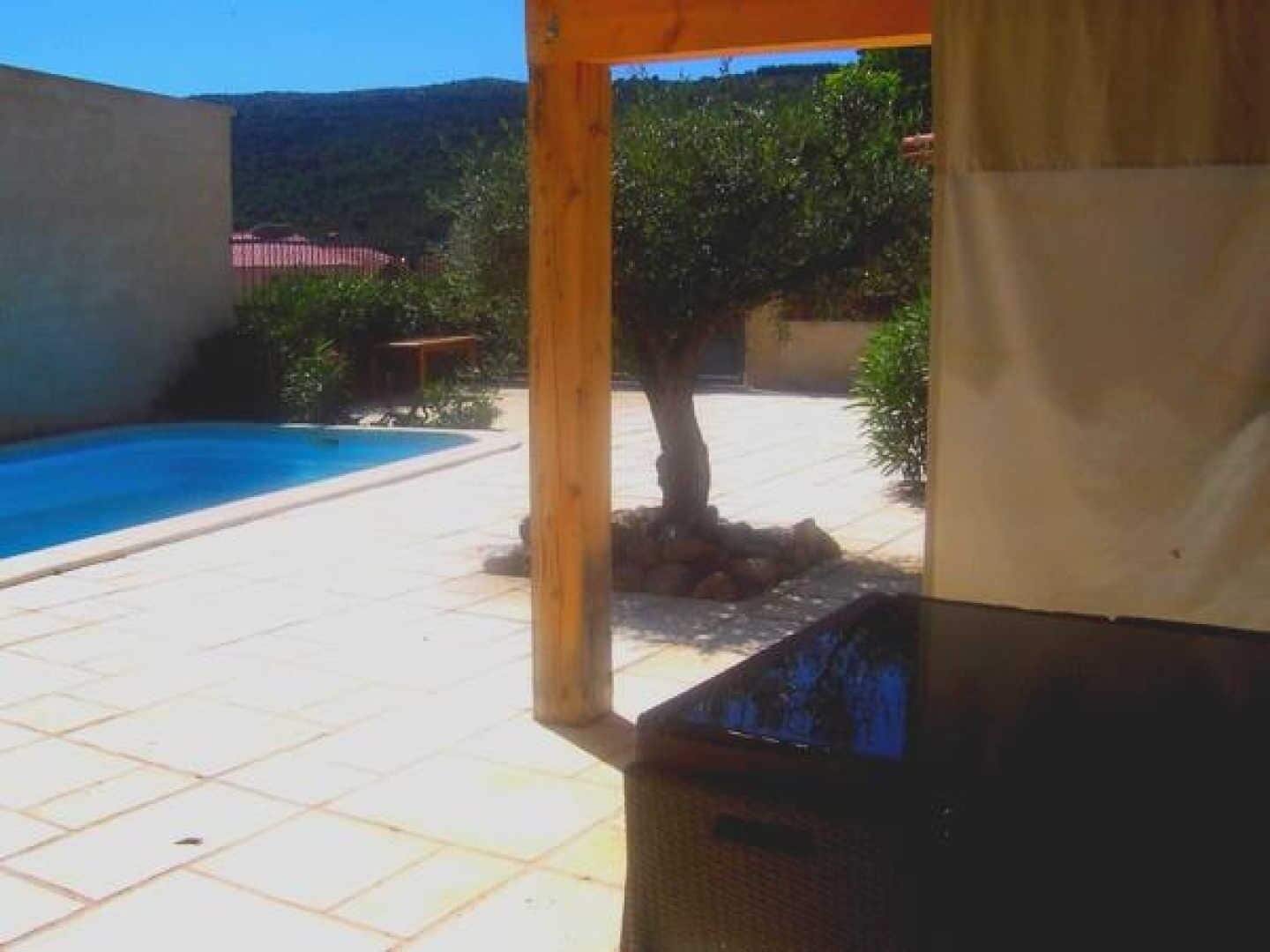 Un área de piscina con suelo de baldosas, una pérgola de madera y un árbol.