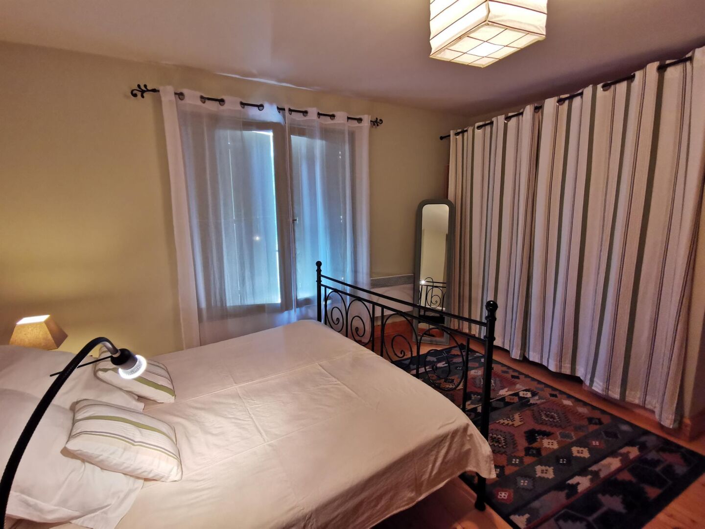 A bedroom with a bed, a bedside lamp, a window with sheer curtains, a mirror, and a striped curtain on the window.
