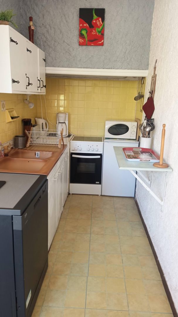 A compact kitchen with a sink, stove, and microwave. There is a small countertop with a kettle and a red tray. The walls are tiled in yellow, and there's a painting of peppers above the stove.