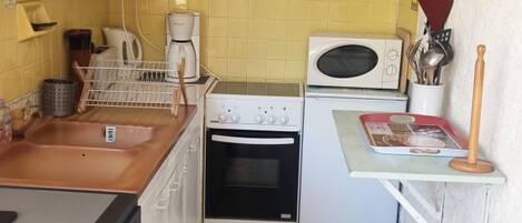 A compact kitchen with a sink, stove, and microwave. There is a small countertop with a kettle and a red tray. The walls are tiled in yellow, and there's a painting of peppers above the stove.