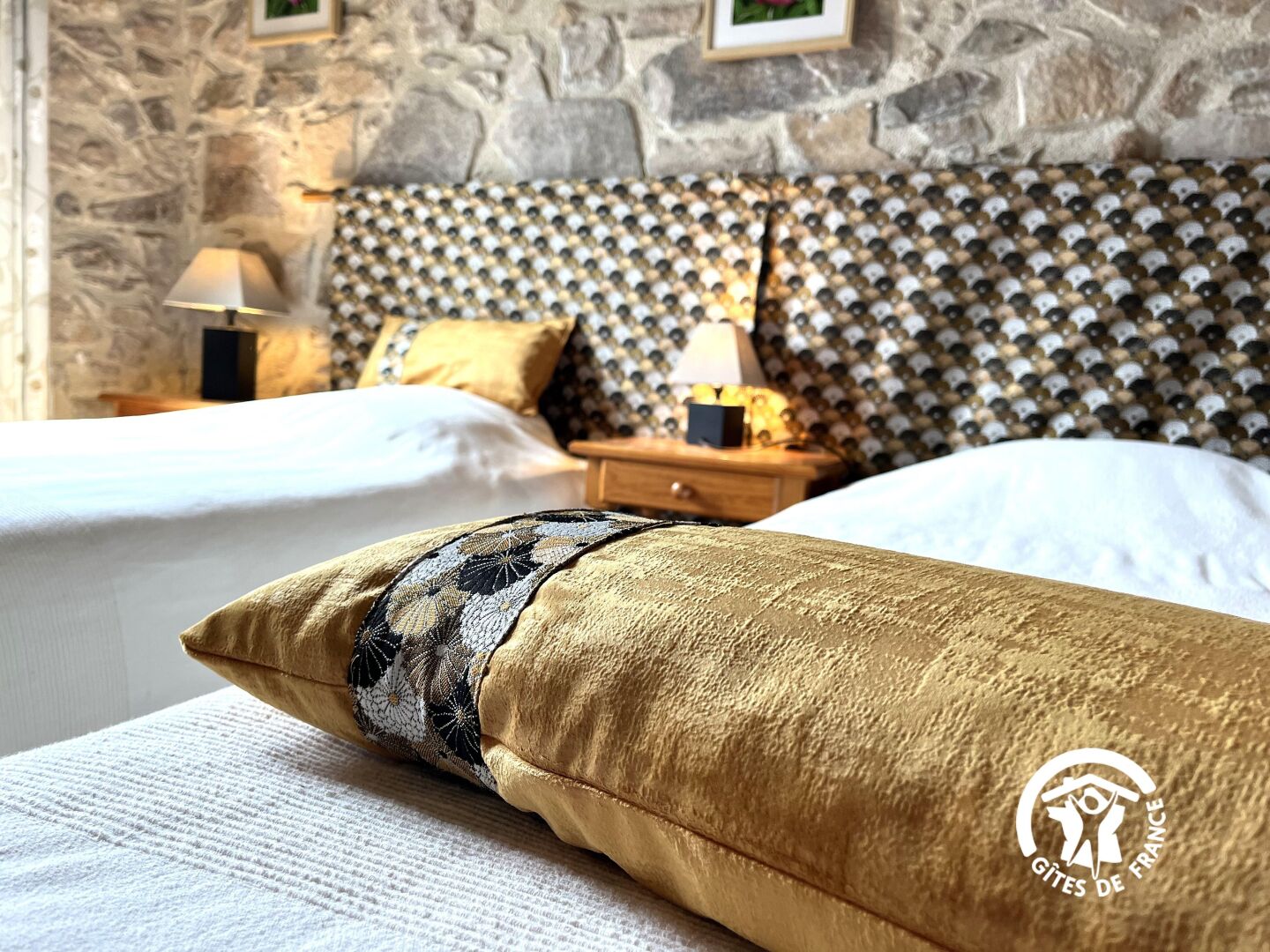 Une chambre d’hôtel avec un mur en pierre, deux lits dotés de têtes de lit à motifs, une table de chevet avec une lampe et un coussin décoratif.