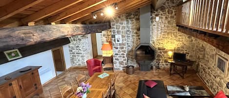 A cozy attic room with a wooden ceiling, stone walls, a fireplace, a dining table with chairs, a sofa, and a window with a wooden railing.