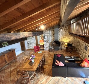 A cozy attic room with a wooden ceiling, stone walls, a fireplace, a dining table with chairs, a sofa, and a window with a wooden railing. - Cal cabre - ST LAURENT DE CERDANS (ST LAURENT DE CERDANS)