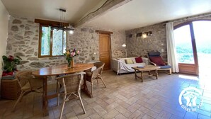 A rustic room with stone walls, a wooden dining table with wicker chairs, a sofa with cushions, and a large window offering a view.