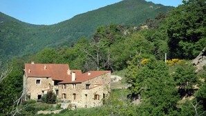 A stone building with a red roof, surrounded by lush greenery and hills in the background.