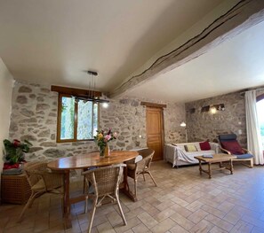 Una habitación rústica con paredes de piedra, una mesa de madera con sillas de mimbre, un sofá con cojines y un gran ventanal con vistas.