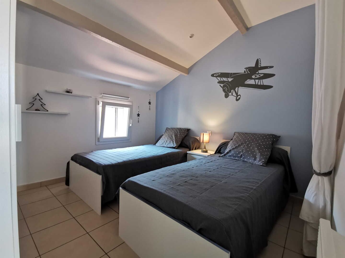 Une chambre avec deux lits simples, une fenêtre, un ventilateur de plafond et une décoration murale représentant un avion.