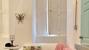 A white sofa with a floral pillow and a pink throw blanket, a small table with a fan and a hat, a patterned rug, and a window with a wooden blind. - Colonial Loft Studio in the best location in the walled city of Ctg. (Cartagena de Indias)