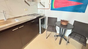 A kitchen with a gas stove, sink, and a painting of a watermelon. There are two wicker chairs and a small table. - Colonial Loft Studio in the best location in the walled city of Ctg. (Cartagena de Indias)