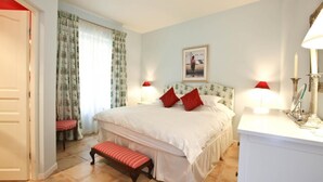 A bedroom with a bed, two bedside lamps, a bench, a bedside table with a mirror, and a wall-mounted picture.
