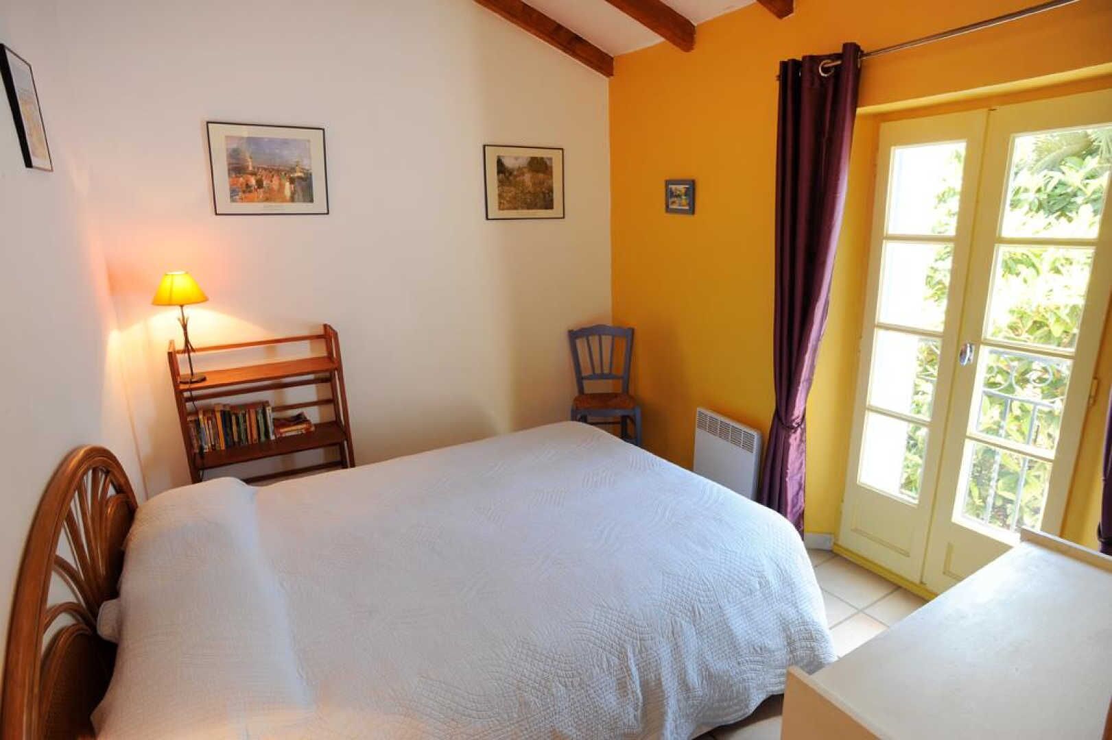 Un dormitorio con cama, una silla, una mesa, una lámpara y una ventana con cortinas.
