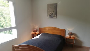 A bedroom with a wooden bed, two bedside tables with lamps, a window with a view of greenery, and a painting on the wall.