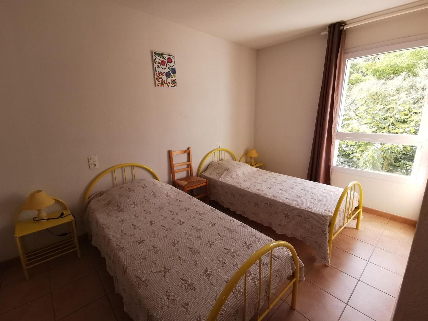 Ein kleiner, schlichter Raum mit zwei Einzelbetten, einem Sessel, einem kleinen Tisch und einem Fenster mit Blick auf Bäume.