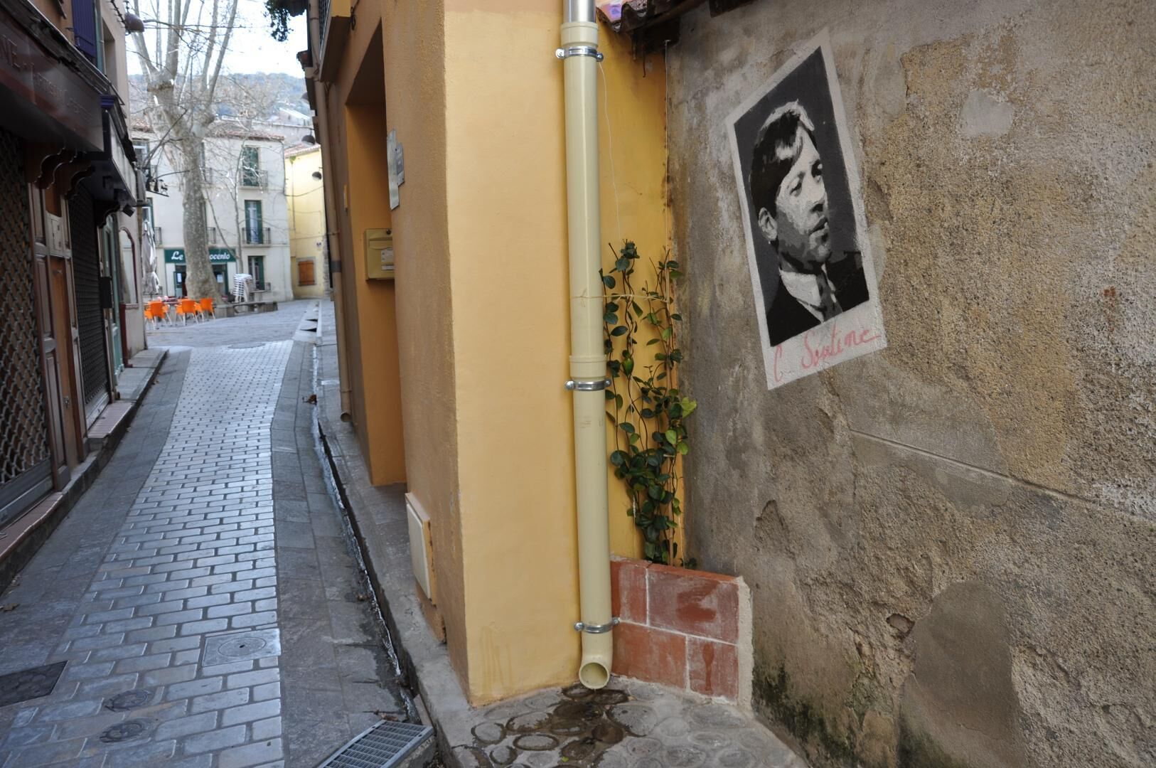 Une rue étroite avec un immeuble jaune, une affiche sur le mur et un tuyau métallique.