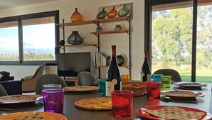 A dining area with a table set for a meal, a television in the background, and a view of the outdoors through large windows. - Eucalyptus - THUIR - Gîte (THUIR)
