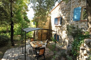 A stone building with a covered outdoor dining area, including a table and chairs, surrounded by trees. - Le mas des cistes - SOREDE (SOREDE)