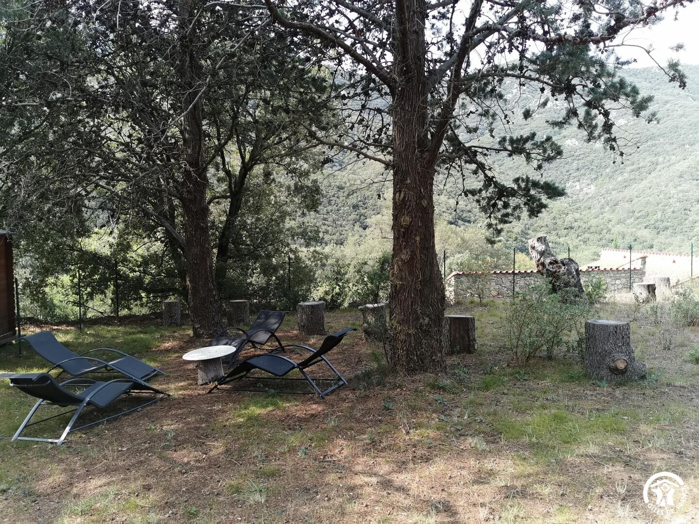 A serene outdoor seating area with lounge chairs and a table, surrounded by trees and a stone wall.