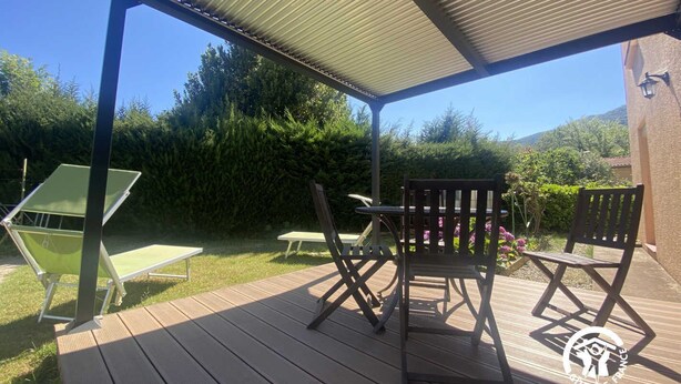 A wooden deck with a covered patio area, outdoor furniture, and a view of a lush garden and trees. - Hortensias - ARLES-SUR-TECH (ARLES-SUR-TECH)