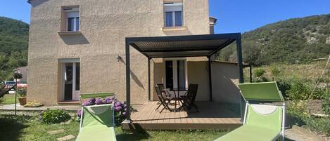 A two-story house with a patio area featuring a table and chairs, surrounded by a garden with purple flowers.