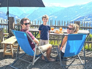 Three people seated at an outdoor table with a mountain view, a wooden fence, and a table with food and drinks.