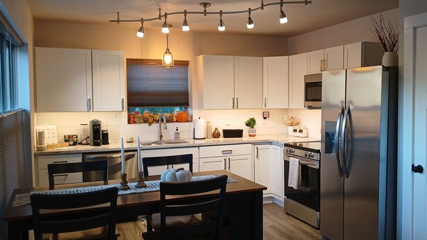 A modern kitchen with white cabinets, a stainless steel refrigerator, and a dining area with a wooden table and chairs. - New charming bungalow with vast mountain views, easy access, stunning sunsets! (Waynesville)