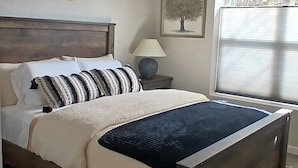 A bedroom with a wooden bed, a ceiling fan, a wall-mounted air vent, a decorative wall hanging, a bedside lamp, and a window with blinds. - New charming bungalow with vast mountain views, easy access, stunning sunsets! (Waynesville)