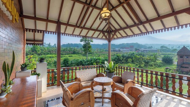 A covered outdoor seating area with wooden furniture and potted plants, overlooking a lush landscape. - Jawa Dwipa Villa (Wonosobo)