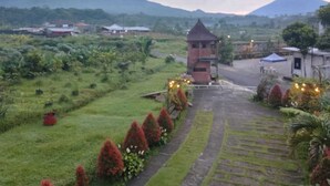 A rural landscape with a small brick structure, possibly a guesthouse, surrounded by lush greenery and a paved path leading to a road. - Jawa Dwipa Villa (Wonosobo)