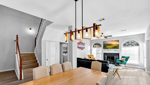 A modern dining area with a wooden table and chairs, a staircase, a living room with a sofa and a fireplace, and a large window with a view. - 5BR Pool House • Just 15 Min to DFW Airport (Lewisville)