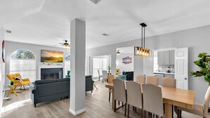 A modern living room with a fireplace, a dining area with a wooden table and chairs, and a kitchen with white cabinets. - 5BR Pool House • Just 15 Min to DFW Airport (Lewisville)