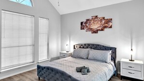 A bedroom with a large bed, a ceiling fan, a wall-mounted artwork, and a nightstand with a lamp. - 5BR Pool House • Just 15 Min to DFW Airport (Lewisville)