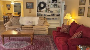 Ein Wohnzimmer mit beiger Couch, roter Couch, Couchtisch aus Holz und einem gemusterten Teppich. Ein Bücherregal mit Dekorationselementen und ein Essbereich mit Stühlen und Tisch.