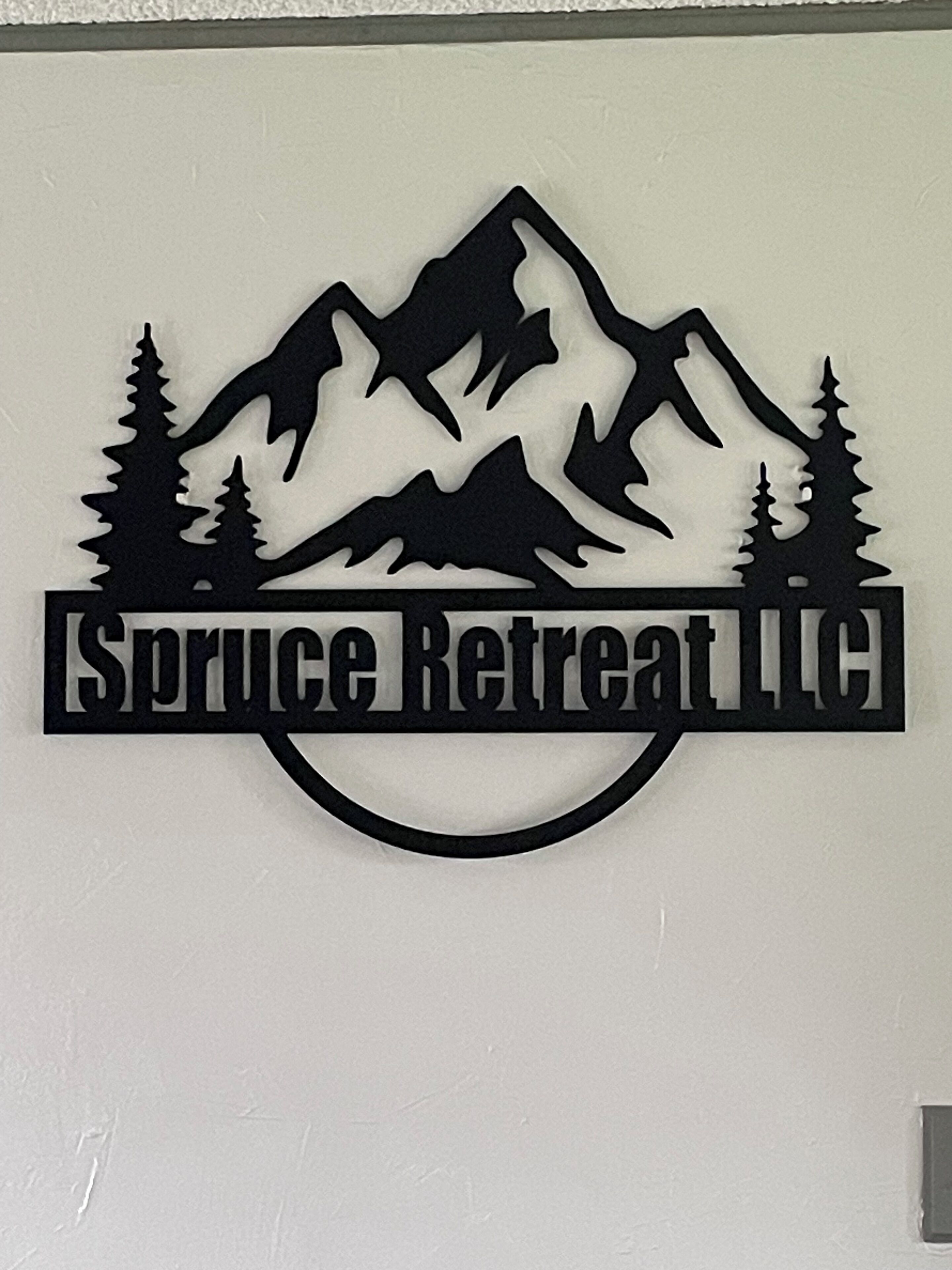 Een wandbord met een berg- en boom silhouet ontwerp, met de tekst 'Spruce Retreat LLC'.