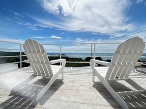 Two white Adirondack chairs on a wooden deck with a view of the ocean and greenery.