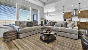 Un salon moderne avec un canapé d’angle, une table basse ronde et une grande fenêtre équipée de stores.