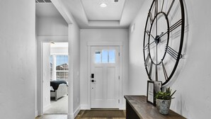 Un couloir avec une grande horloge murale, un banc en bois agrémenté d’une plante en pot et une porte donnant sur une chambre.