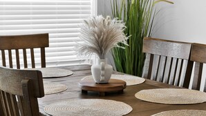Une table à manger ornée d’un vase blanc contenant des herbes de la pampa séchées, entourée de sets de table en fibres tressées et de chaises en bois.