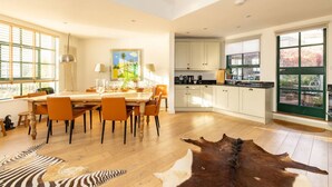 A spacious kitchen with white cabinets, a dining area with a wooden table and orange chairs, and a large window with blinds.