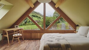 Ein Schlafzimmer mit großem Fenster, Holzbalken, einem Bett, einem Sessel und einem Schreibtisch.